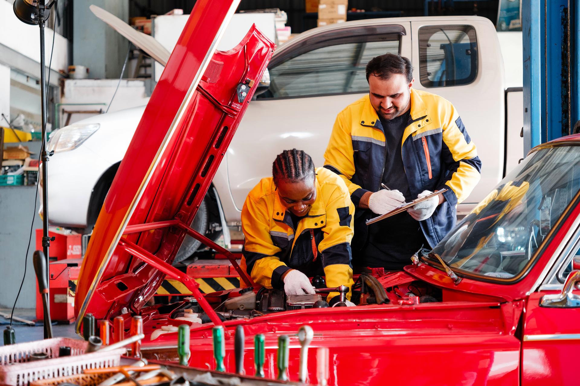 African woman and an Indian man, both professional auto mechanics, work together in busy garage. Woman repairs the red car's engine while man takes notes. Teamwork and diversity in automotive industry