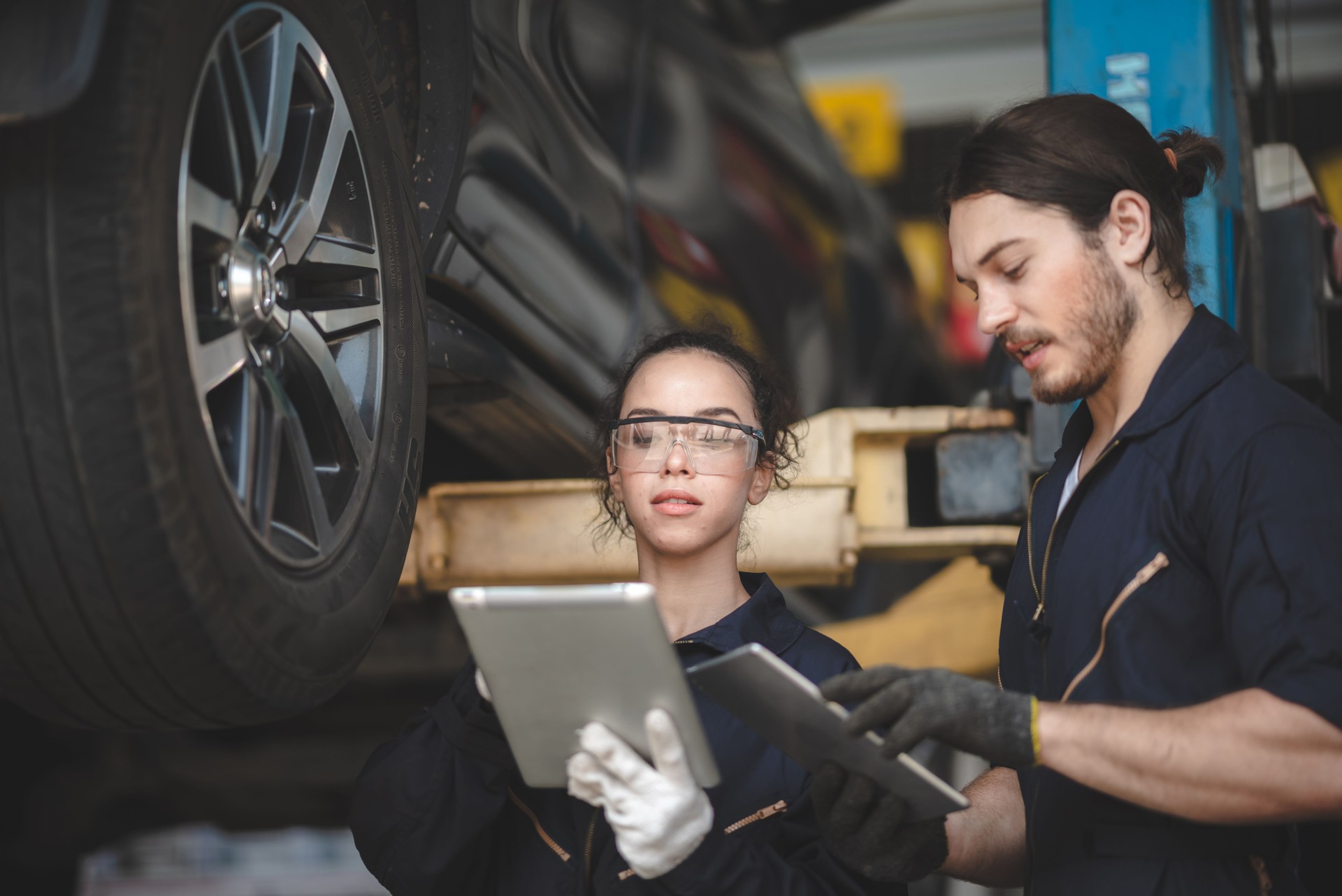 Young male and female car mechanic wearing uniform with safety eye glasses and hand gloves working on digital tablet while diagnosing problem in car
