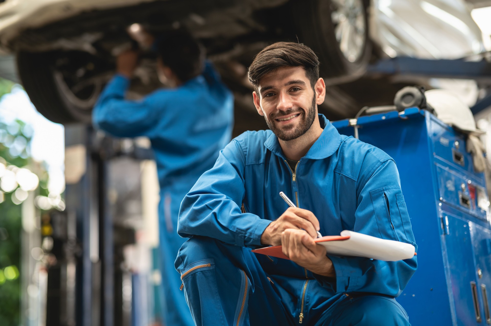 Portrait photo of professional look Caucasian vehicle service technician kneeling in front of car repair shop and holding car inspect check list.